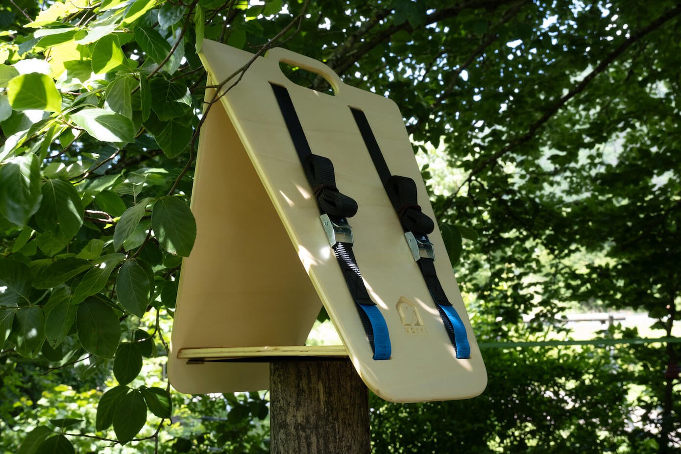 View of a triangular wooden bird feeder by Ulap design sitting on a pole in the middle of a grove. Products from the MÖKKI collection help promote biodiversity.