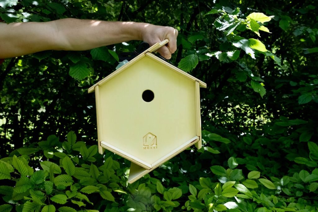 View of a hexagonal wooden bird house by Ulap design held in the hand of a man. You can see the arm holding the birdhouse in front of the trees. Products from the MÖKKI collection help promote biodiversity.