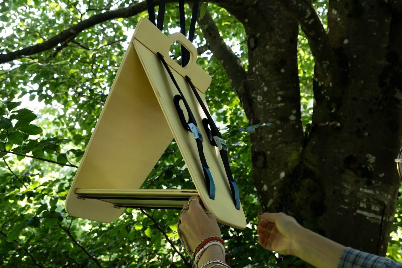 Close-up view of a triangular wooden bird feeder by Ulap design hanging on the branch of a beech tree. You can see the hands of a woman installing the feeder in the best position to attract local wildlife. Products from the MÖKKI collection help promote biodiversity.