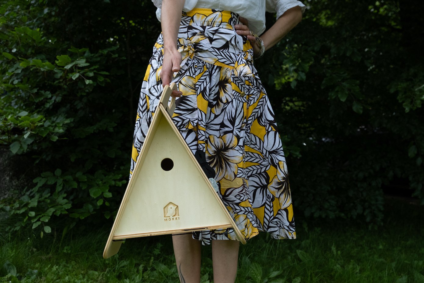View of a triangular wooden birdhouse of Ulap design held in the hand by a woman with a large yellow and brown colored skirt standing barefoot in the middle of a forest. Products from the MÖKKI collection help promote biodiversity.