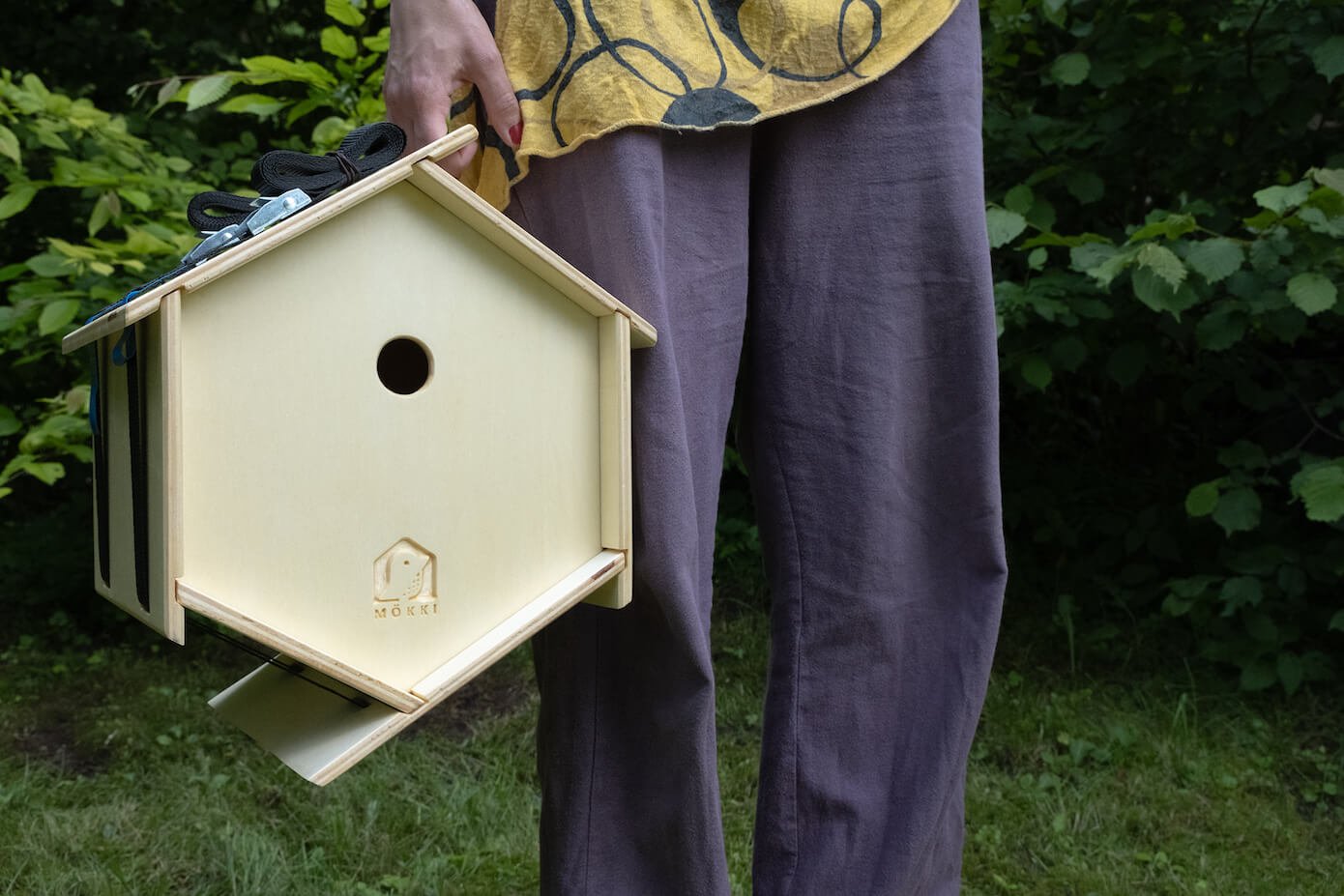 View of a hexagonal wooden bird house by Ulap design held in the hand of a woman. You can see the woman standing, facing the trees of a forest, holding the bird feeder by the handle. Products from the MÖKKI collection help promote biodiversity.