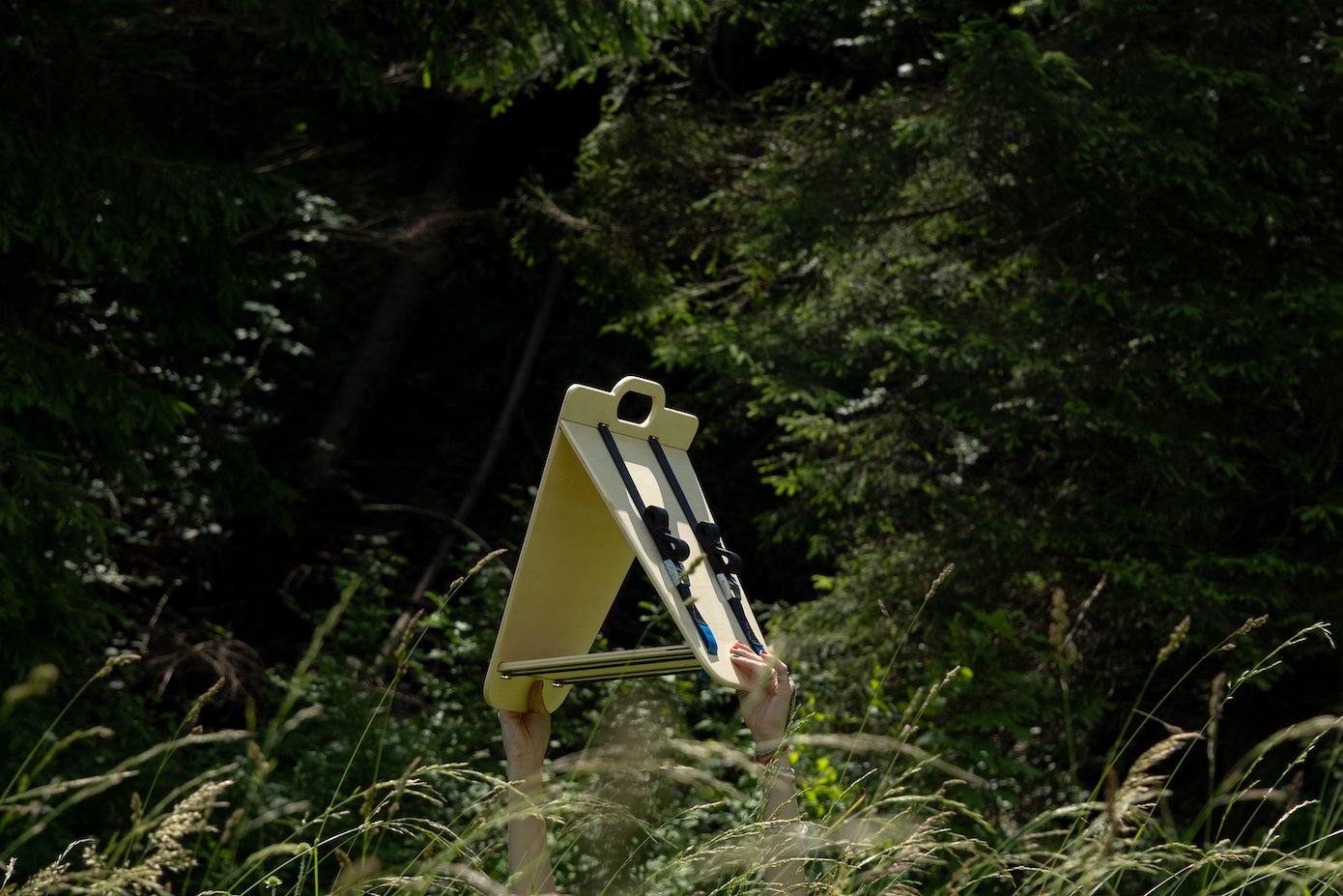 View of a triangular wooden bird feeder by Ulap design held in the hand of a woman. You can see the two arms holding the bird feeder emerge from the grass and flowers. Products from the MÖKKI collection help promote biodiversity.