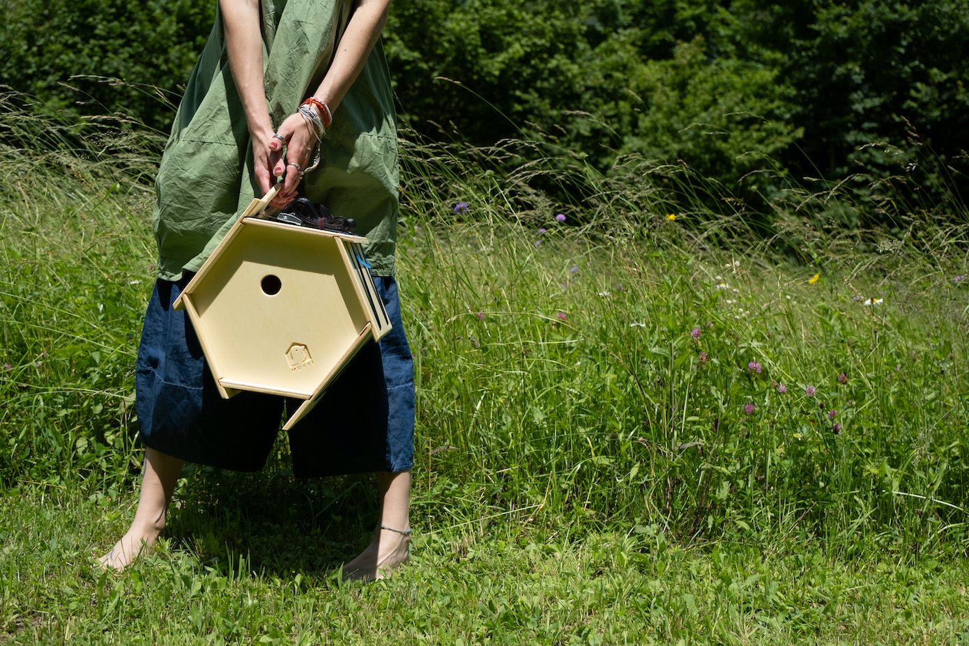 View of a hexagonal wooden birdhouse by Ulap design held in the hand by a barefoot woman amidst a flowery meadow. Products from the MÖKKI collection help promote biodiversity.