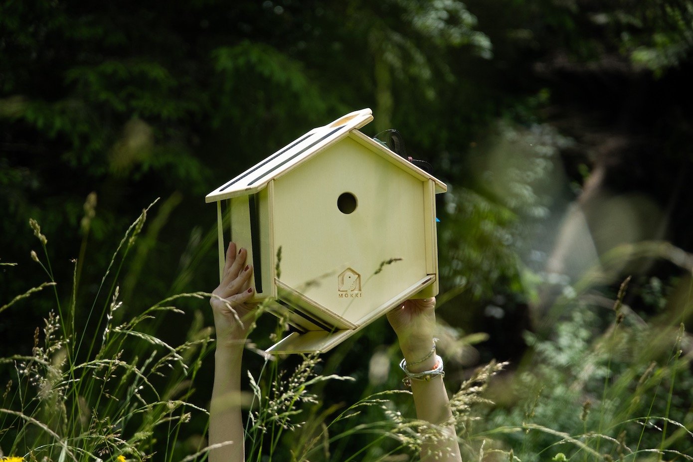 View of a hexagonal wooden bird house by Ulap design held in the hand of a woman. You can see the two arms holding the birdhouse emerge from the grass and flowers. Products from the MÖKKI collection help promote biodiversity.