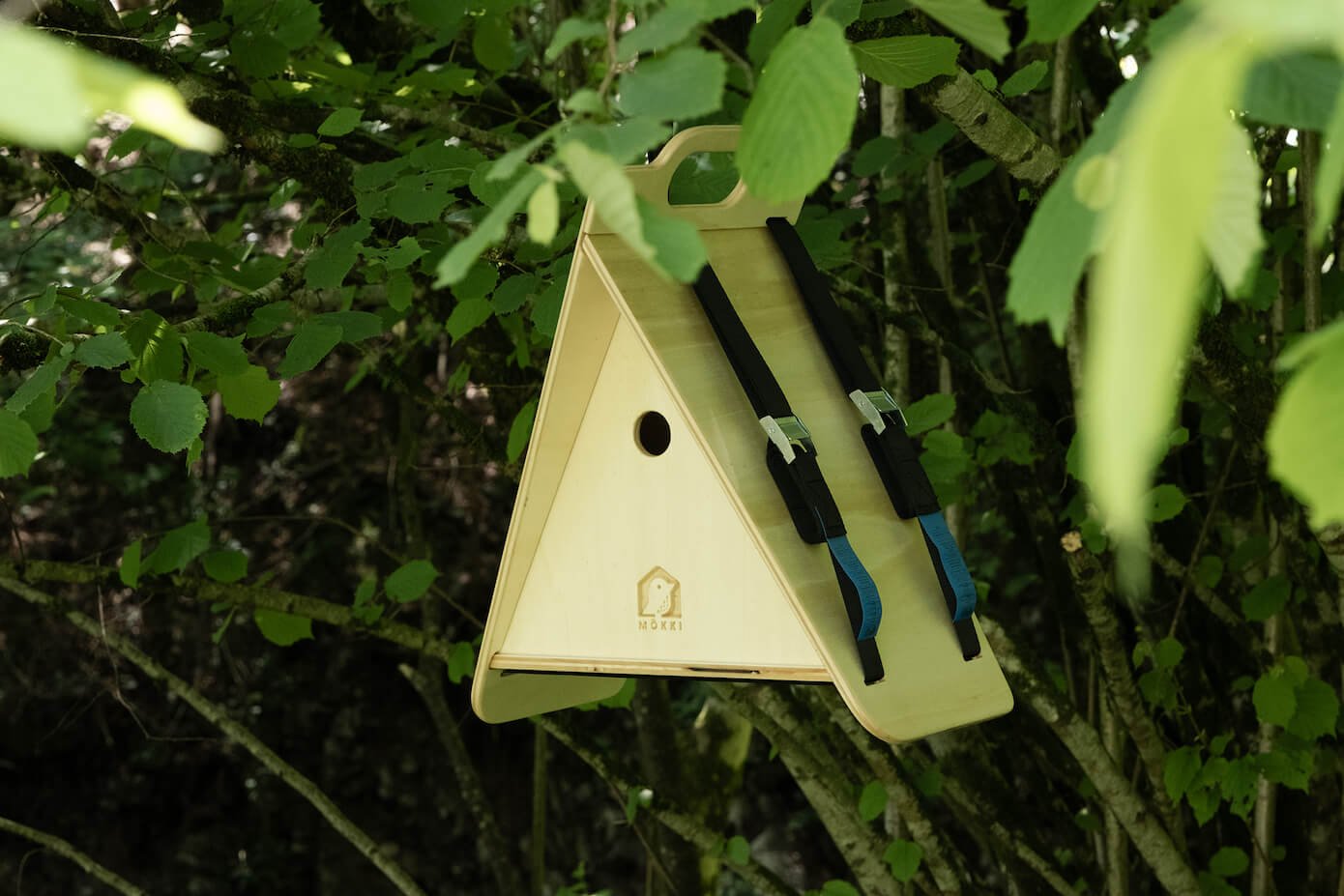View of a triangular wooden birdhouse by Ulap design hanging from a hazel tree. Products from the MÖKKI collection help promote biodiversity.