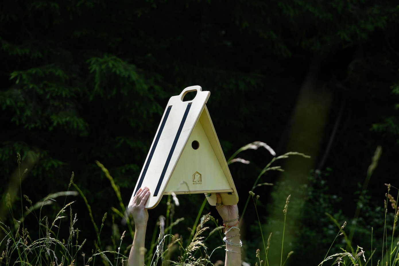 View of a triangular wooden bird house by Ulap design held in the hand of a woman. You can see the two arms holding the birdhouse emerge from the grass and flowers. Products from the MÖKKI collection help promote biodiversity.