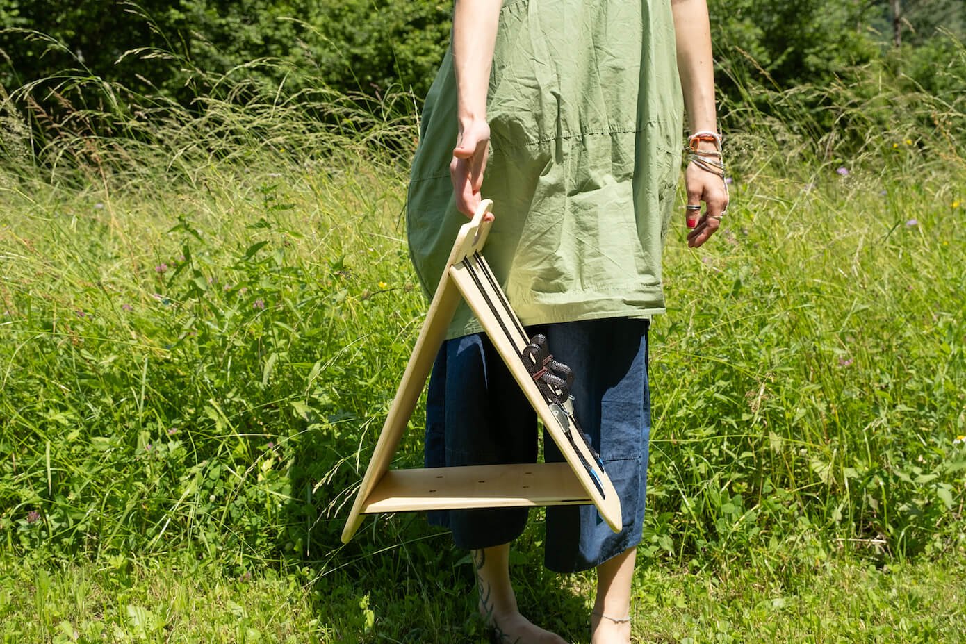 Frontal view of a triangular wooden bird feeder by Ulap design held in the hand of a woman. You can see the woman standing in a meadow with wild flowers, holding the bird feeder by the handle. Products from the MÖKKI collection help promote biodiversity.