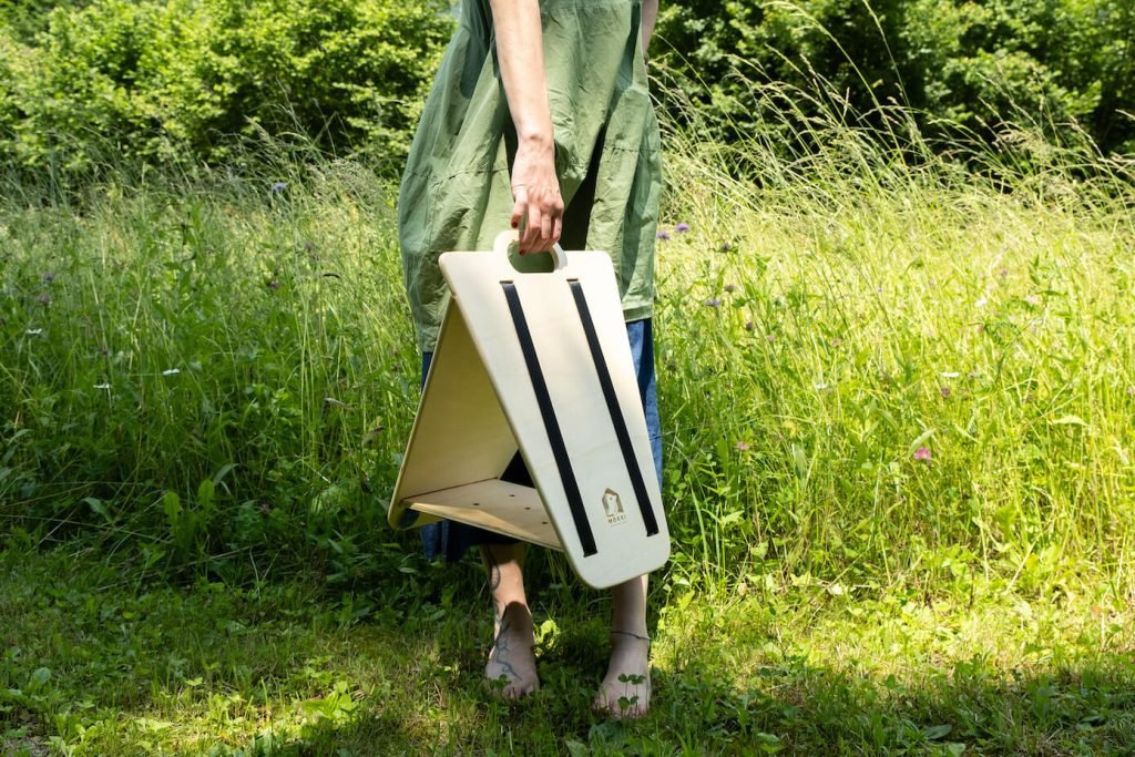 View of a triangular wooden bird feeder by Ulap design held in the hand of a woman. You can see the woman standing in a meadow with wild flowers, holding the bird feeder by the handle. Products from the MÖKKI collection help promote biodiversity.